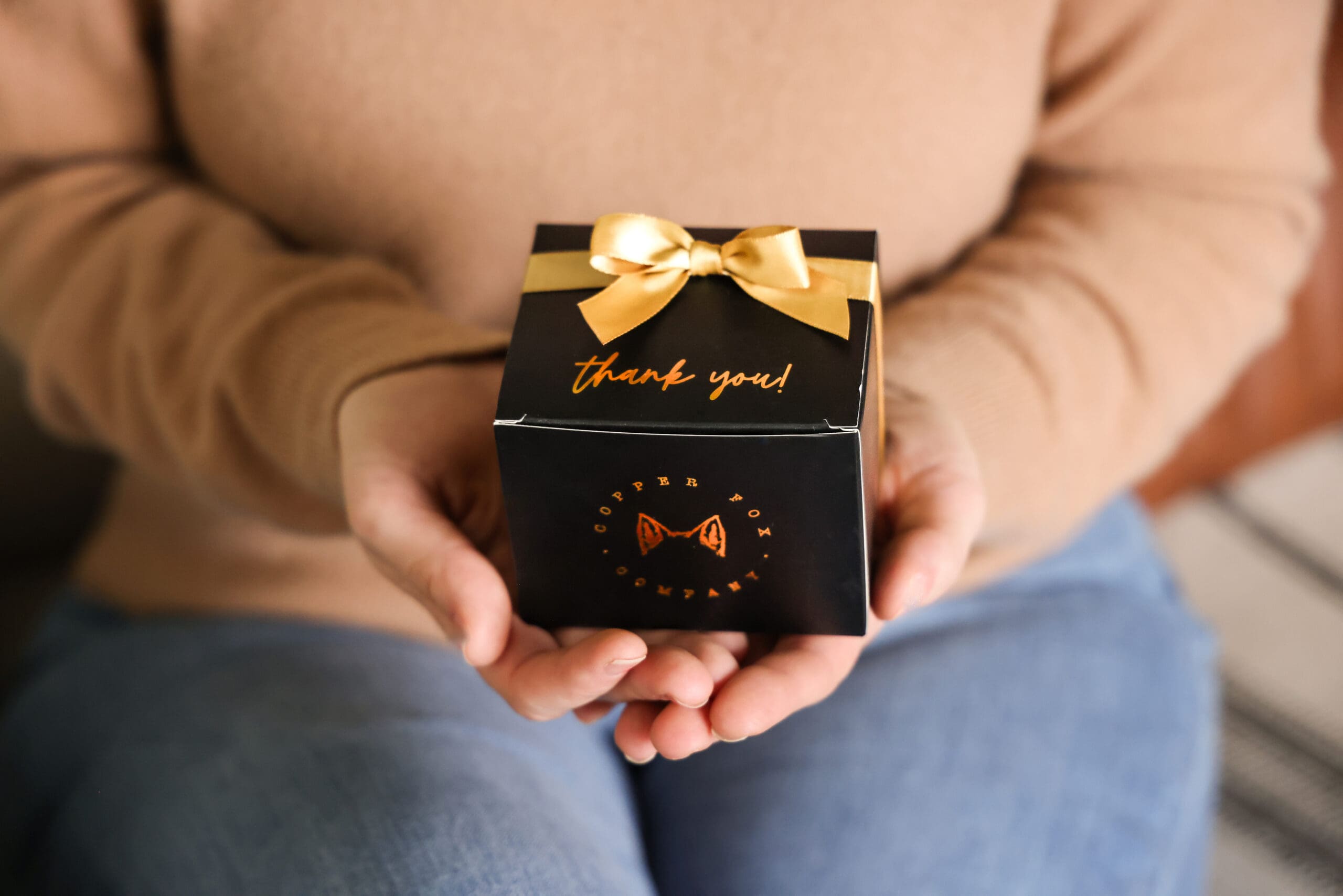 A person sitting and holding an elegantly wrapped gift box with a ribbon bow and foil script branding, illustrating the transition from a standard delivery to a curated customer experience moment.