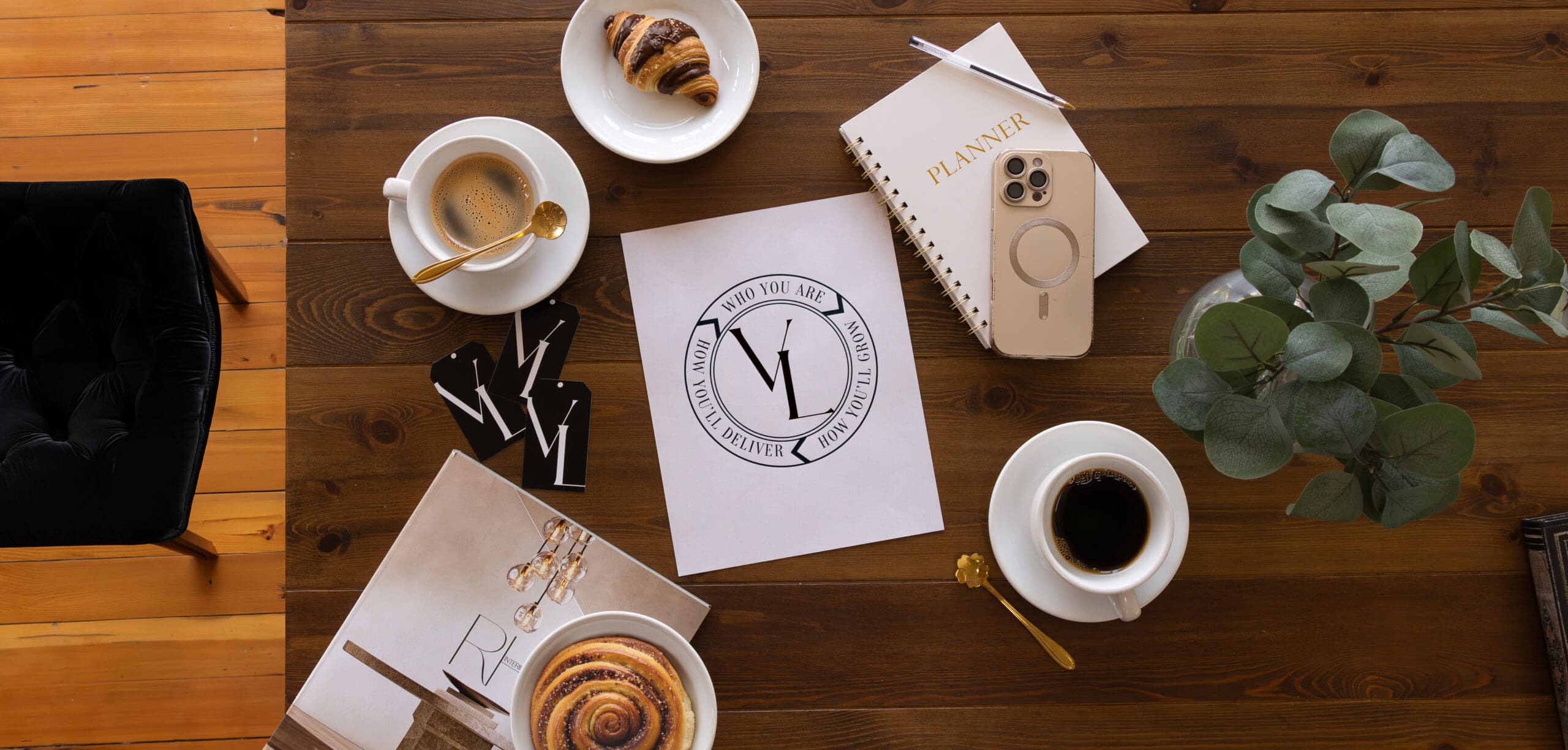 A curated studio workspace featuring a wooden table with a warm cup of coffee and a stack of books. In the center, a printed graphic illustrates The Velvet Circle, a three-part continuous loop showing the relationship between brand identity, growth strategy, and client delivery.