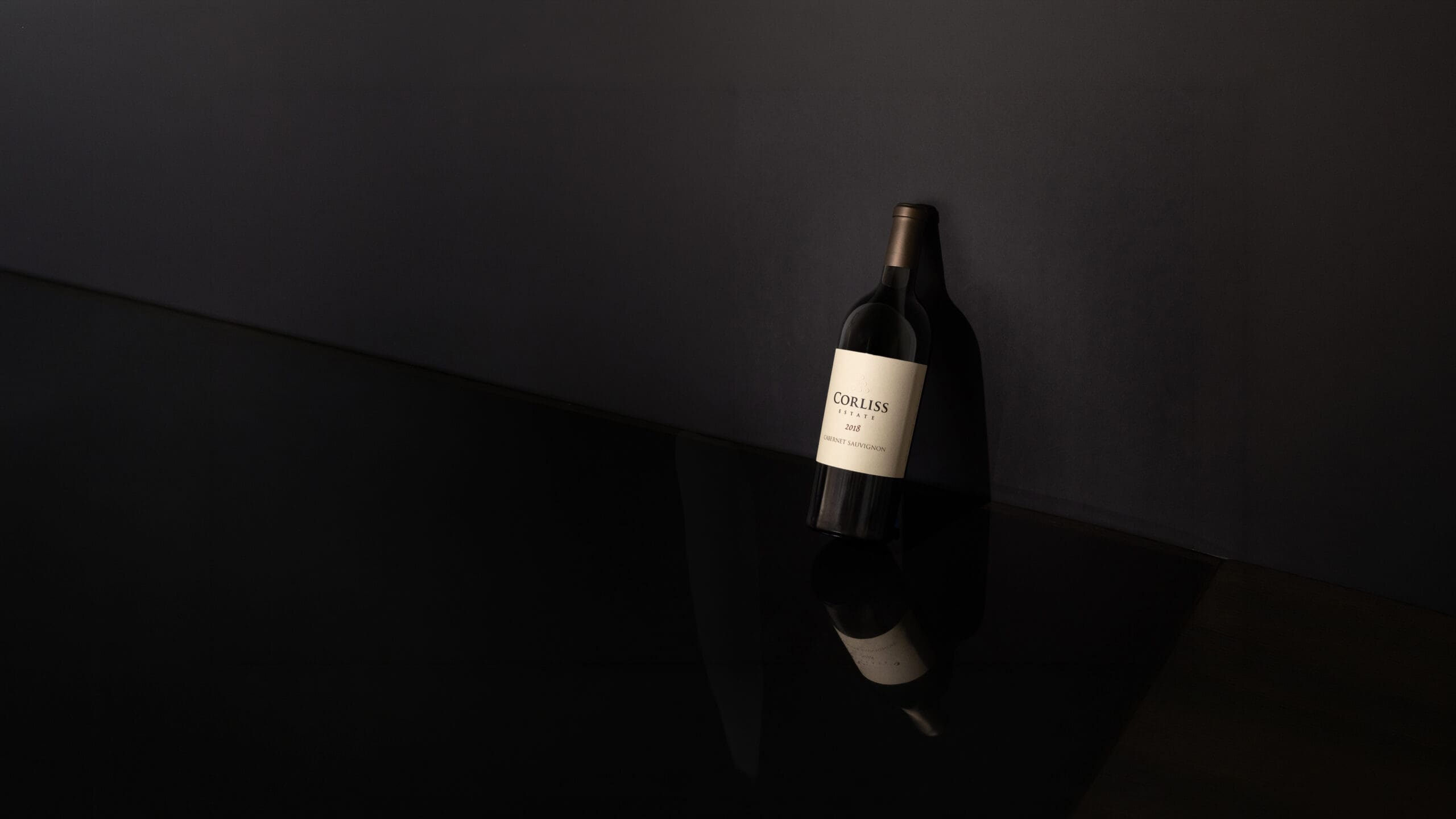 A dark, moody product photo of a wine bottle with a white label, standing on reflective black glass against a matte black background.
