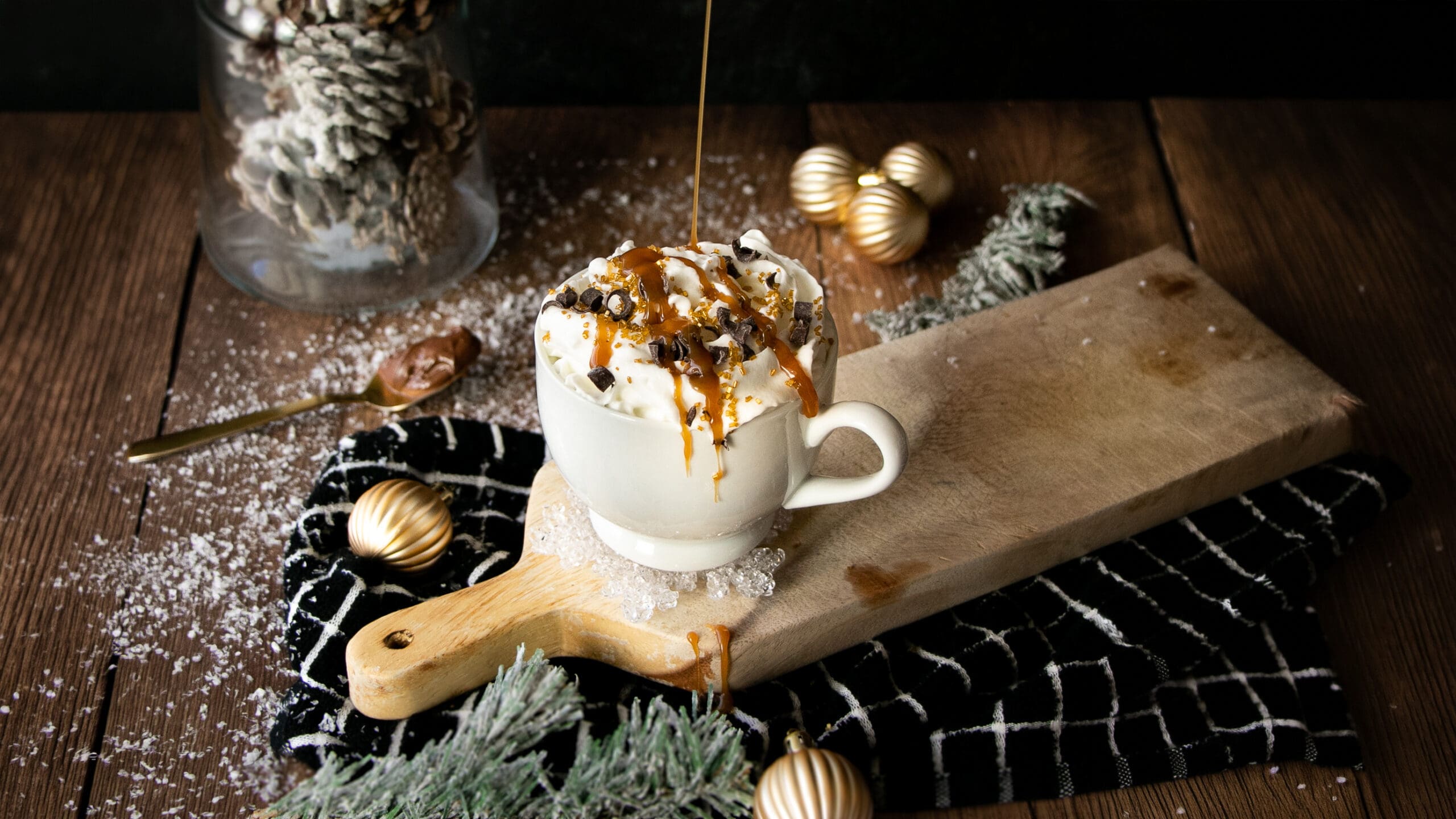 A cozy winter scene featuring a gourmet hot chocolate with whipped cream, caramel drizzle, and chocolate curls on a rustic wooden table with pine sprigs and snow.