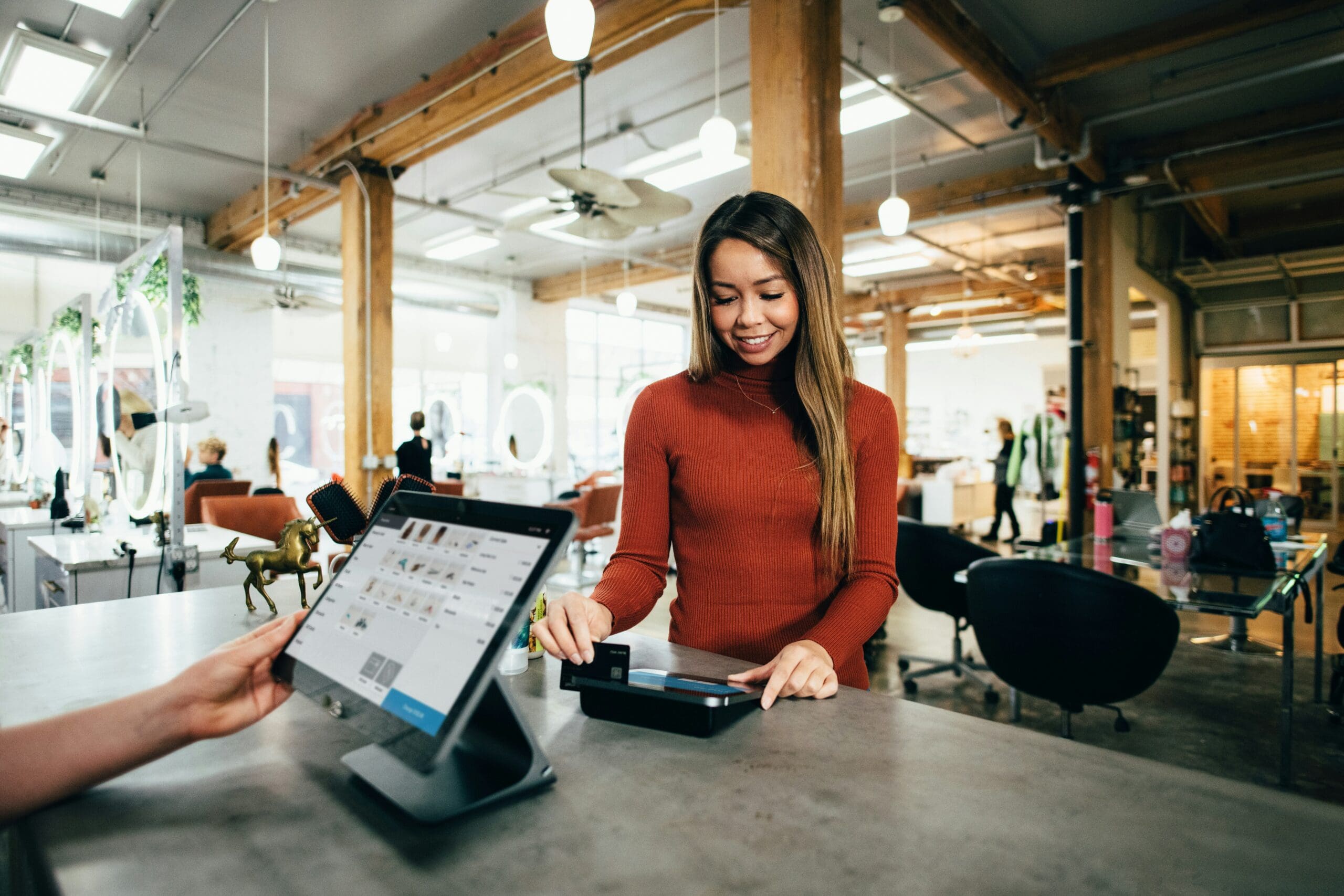 A female customer smiling warmly while interacting with a business owner at a wooden service counter in a sunlit, boutique store with rustic wood beams.